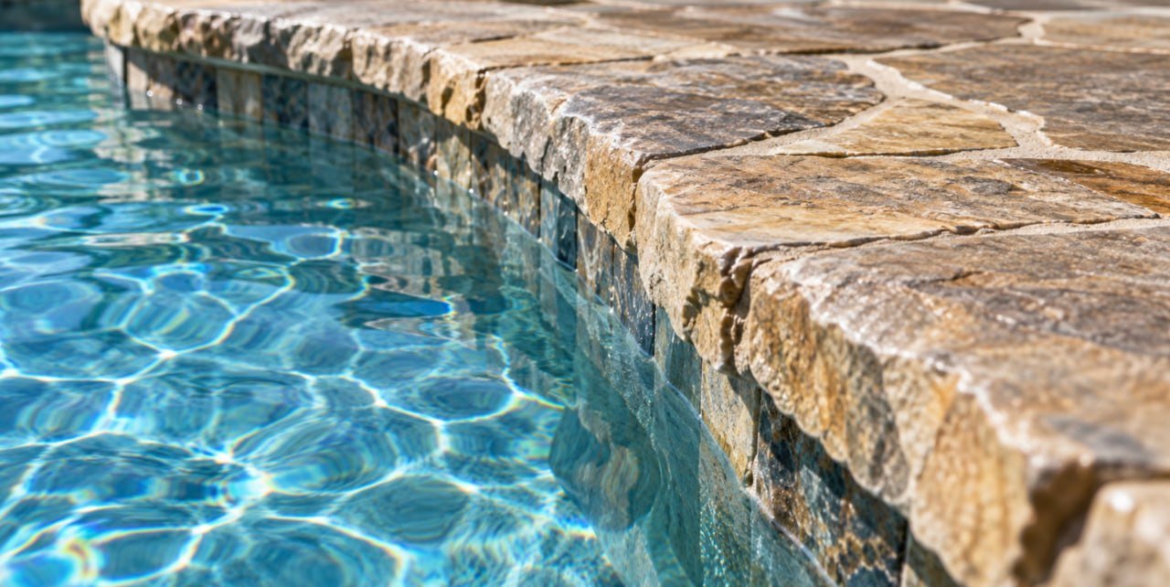 A curved pool edge with irregular stone coping above a dark tile band