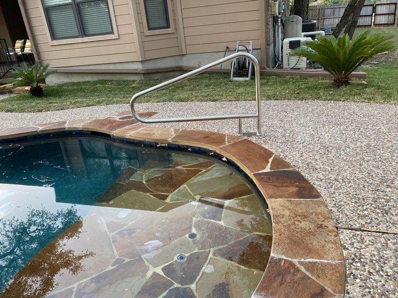 Curved stone entry steps under clear water, with a stainless handrail and pebble deck
