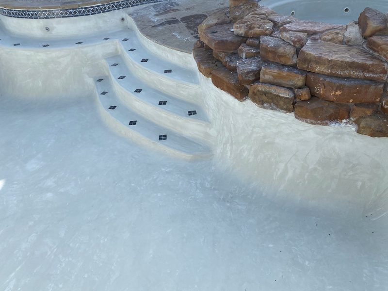 An empty pool with fresh pale plaster, curved entry steps and dark tile accents beside a large rock feature
