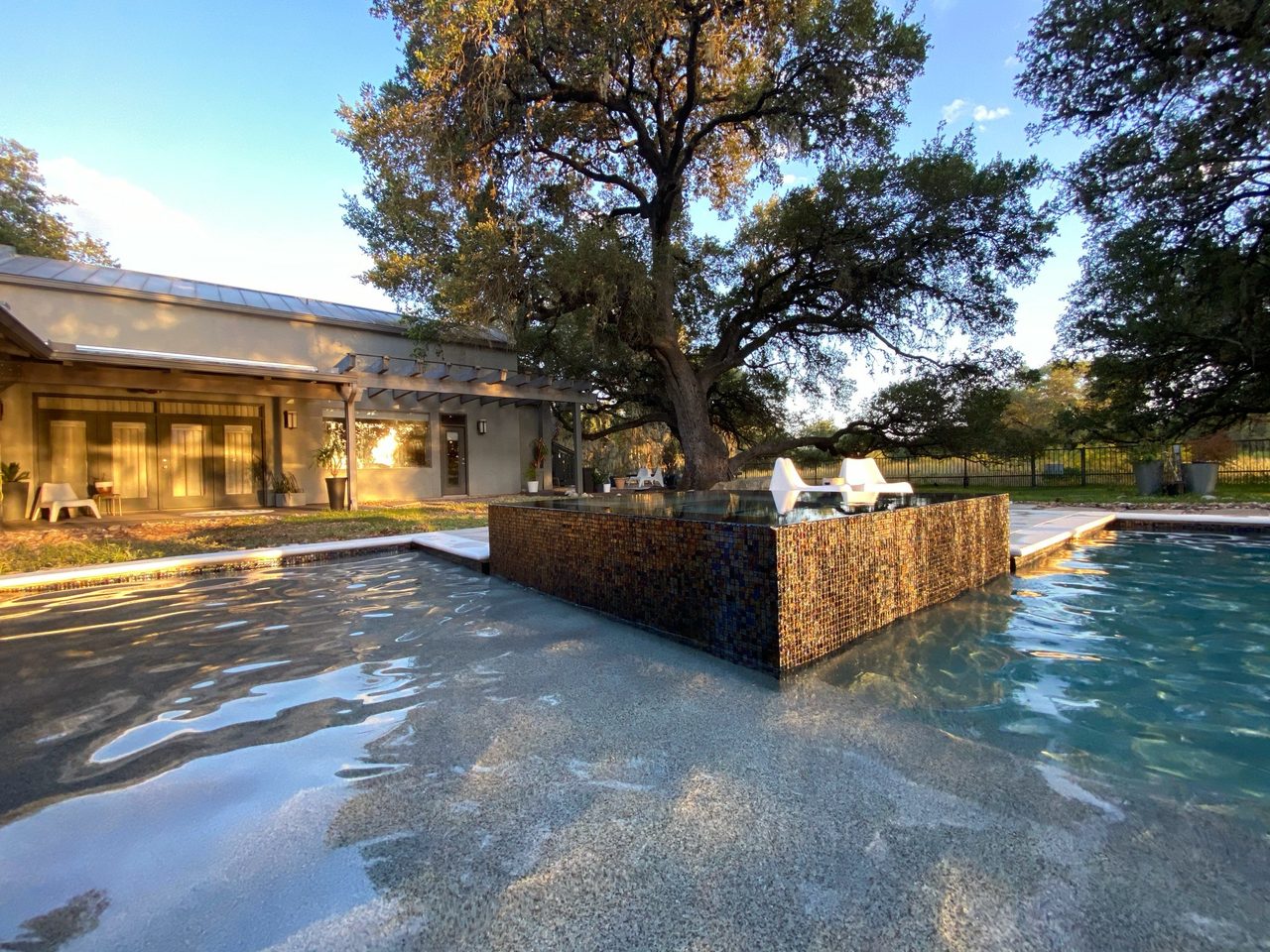 A modern pool seen from the waterline: a shallow shelf, dark-tiled raised feature and white loungers in warm light