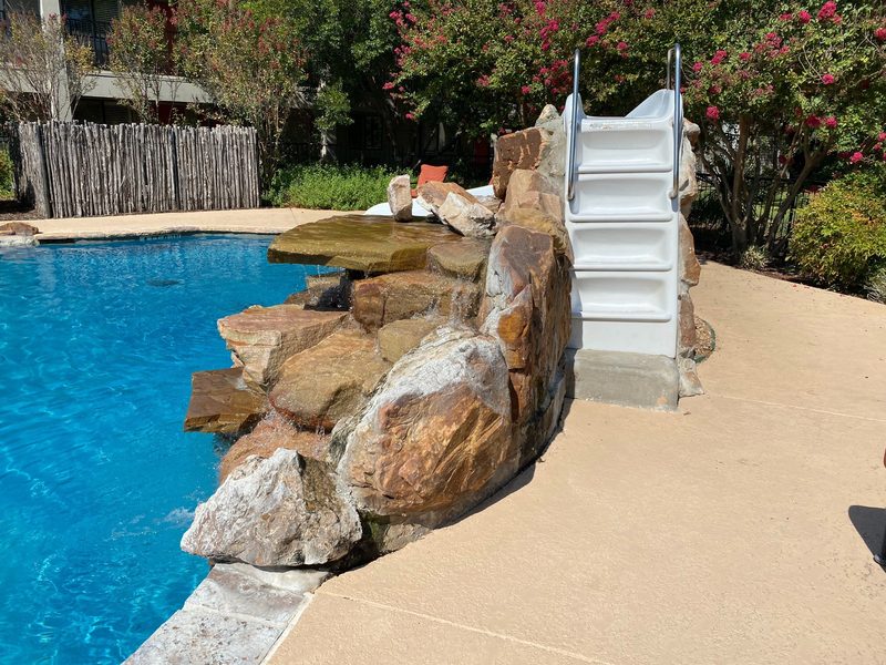 A blue pool beside a rock waterfall, with white stairs and rails up to a slide