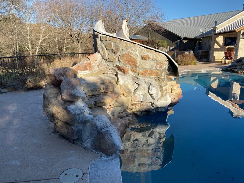 The stone enclosure behind a pool slide, calm water and a concrete deck beside it