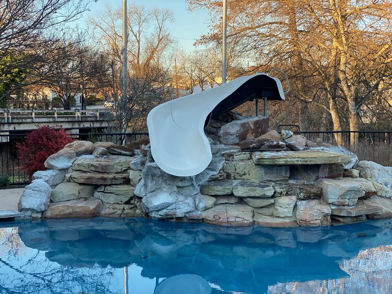 A white curved slide over a broad stacked-stone waterfall, still water in front
